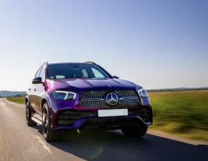A purple Mercedes-Benz SUV driving along a rural road on a sunny day, with green fields and blue sky in the background.
