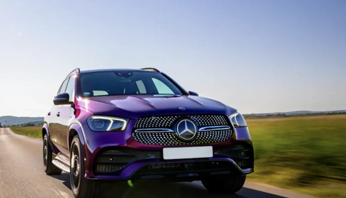 A purple Mercedes-Benz SUV driving along a rural road on a sunny day, with green fields and blue sky in the background.