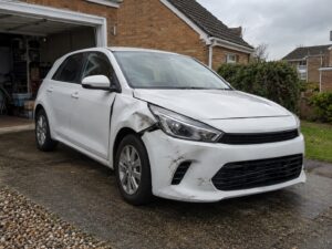 A white hatchback with front wing and bumper damage parked on a British house driveway with the garage door open.