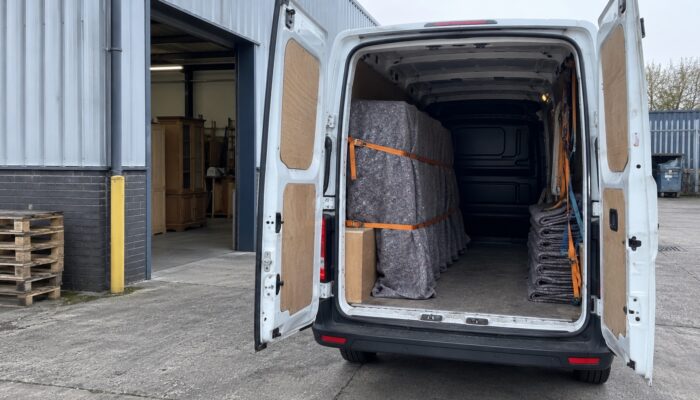 The open back of a delivery van at a furniture shop's loading door, moving blankets and straps inside and a wrapped item strapped in place