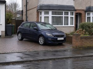 A dark blue family hatchback parked on the block paved driveway of a British suburban house on an overcast day.