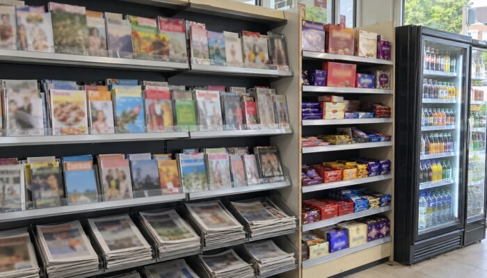 Magazine and newspaper racking, a shelf of boxed confectionery and a glass-fronted drinks chiller in a newsagent