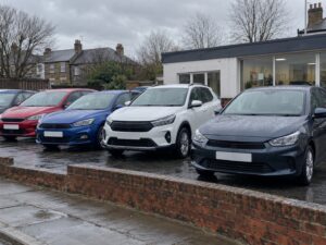Used cars of different colours parked on the forecourt of a small British car dealership on a wet, overcast day.